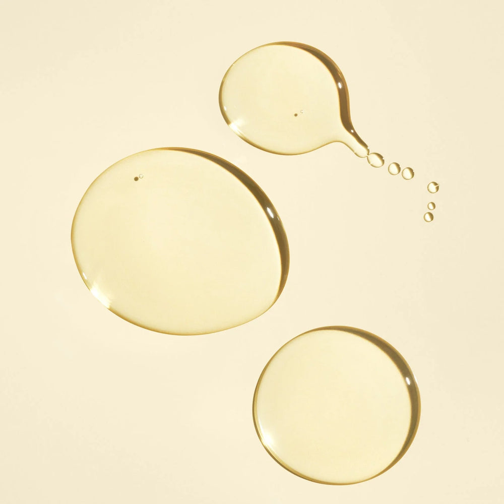 Texture dorée et translucide de l'huile capillaire nourrissante ORIBE Gold Lust, présentée en trois gouttes brillantes de différentes tailles sur un fond beige crème.