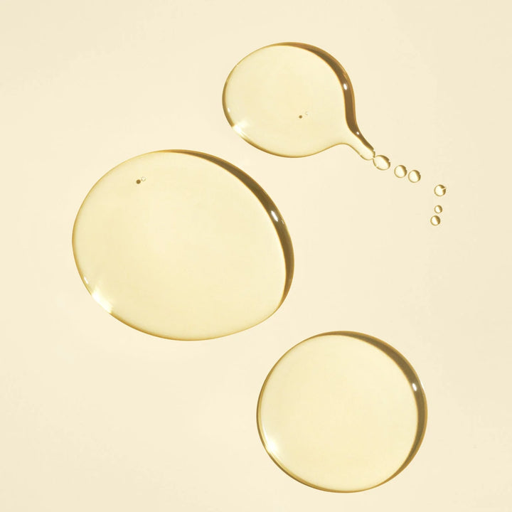 Texture dorée et translucide de l'huile capillaire nourrissante ORIBE Gold Lust, présentée en trois gouttes brillantes de différentes tailles sur un fond beige crème.