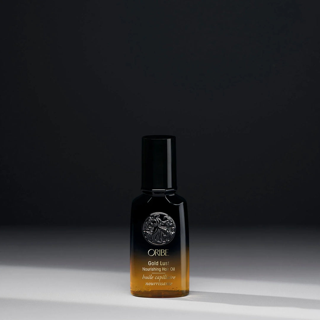 Huile capillaire nourrissante ORIBE Gold Lust 50 ml format voyage, flacon noir dégradé ambré avec emblème argenté et bouchon noir, photographié sur fond sombre.
