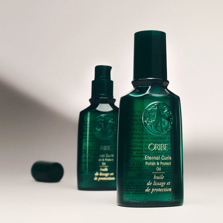 Huile capillaire Oribe Eternal Curls Polish & Protect Oil avec bouteille au premier plan, seconde bouteille floutée et capuchon posé à côté sur fond clair.