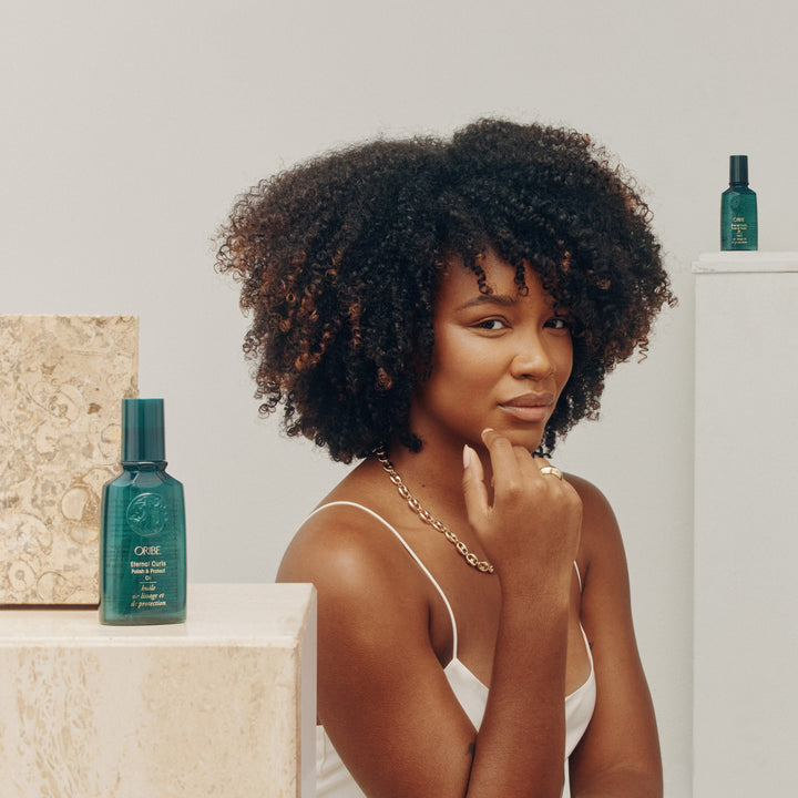 Femme aux cheveux bouclés volumineux avec boucles brillantes devant un décor épuré, accompagnée de l’huile Oribe Eternal Curls Polish & Protect Oil placée sur des blocs décoratifs en arrière-plan.