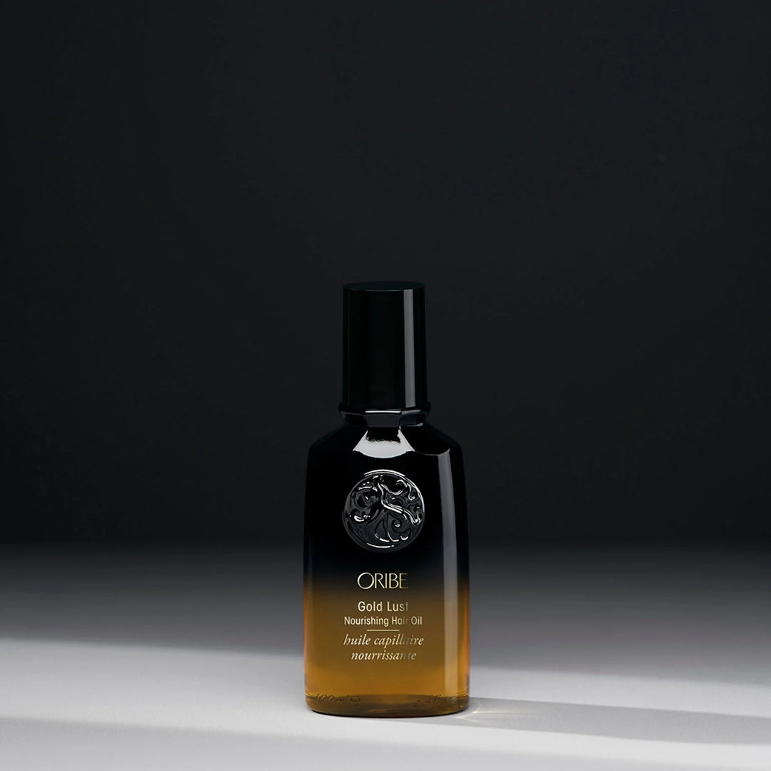 Huile capillaire nourrissante ORIBE Gold Lust 100 ml, flacon en verre dégradé noir et ambré avec bouchon noir brillant, emblème argenté et inscriptions dorées, sur fond sombre.