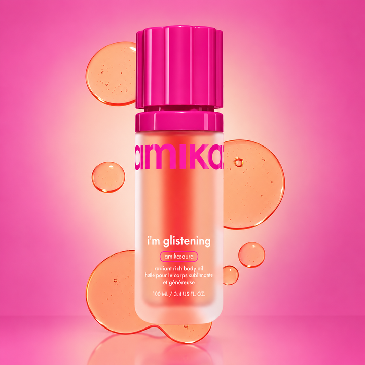 Huile pour le corps AMIKA I'm Glistening 100 ml en flacon rose, avec gouttes d'huile orangée sur fond rose et caractéristiques de la formule.