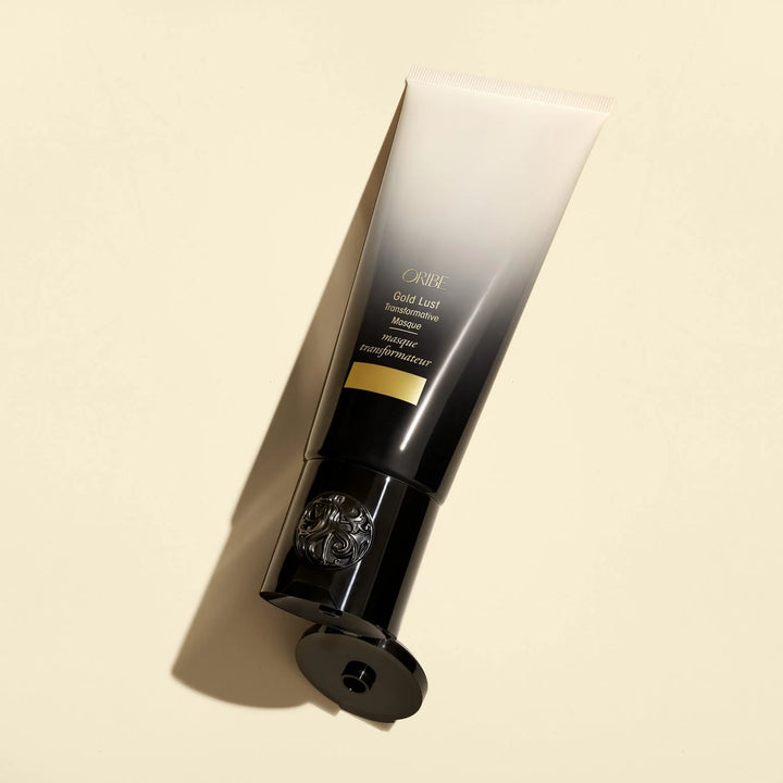 Masque transformateur ORIBE Gold Lust 150 ml, tube dégradé ivoire et noir avec détails dorés et bouchon noir ouvert, posé en diagonale sur un fond beige clair.
