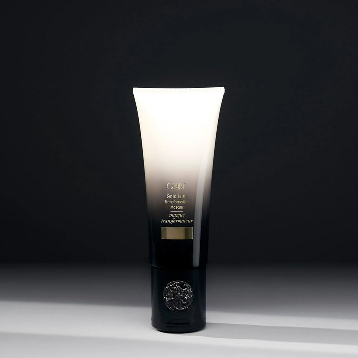Masque transformateur réparateur ORIBE Gold Lust 150 ml, tube dégradé blanc ivoire et noir avec inscriptions dorées, bouchon noir orné d'un emblème argenté, sur fond sombre.