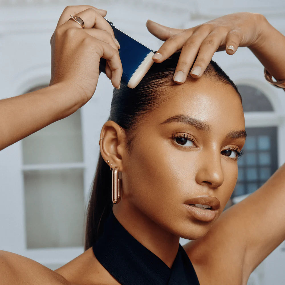 Femme appliquant le bâton de cire à lisser ORIBE Supershine sur une coiffure sleek pour discipliner les frisottis et lisser les cheveux.