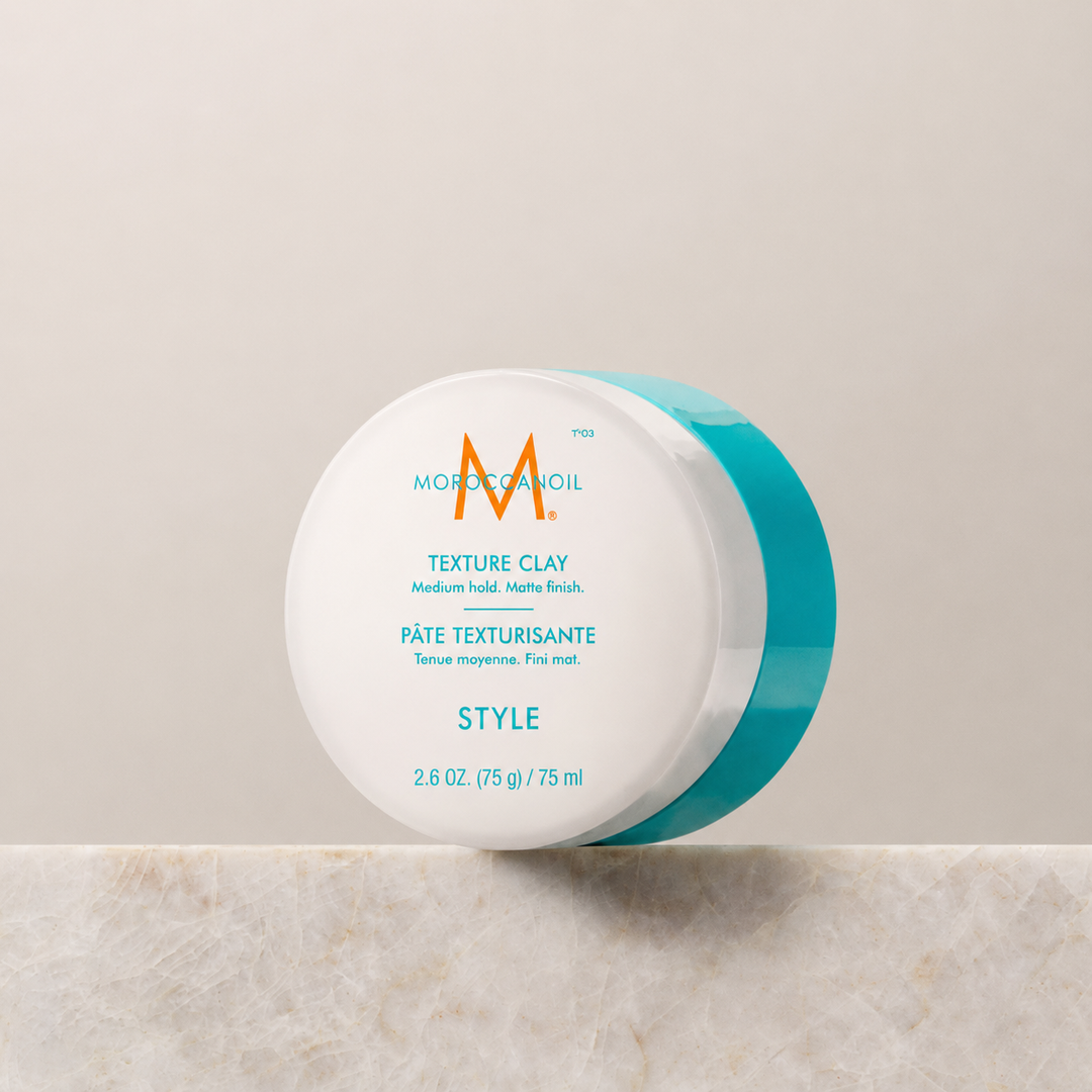 Pâte texturisante Moroccanoil Style 75 ml en pot blanc avec couvercle turquoise, posée sur un support en pierre, fond beige minimaliste, photo produit professionnelle.