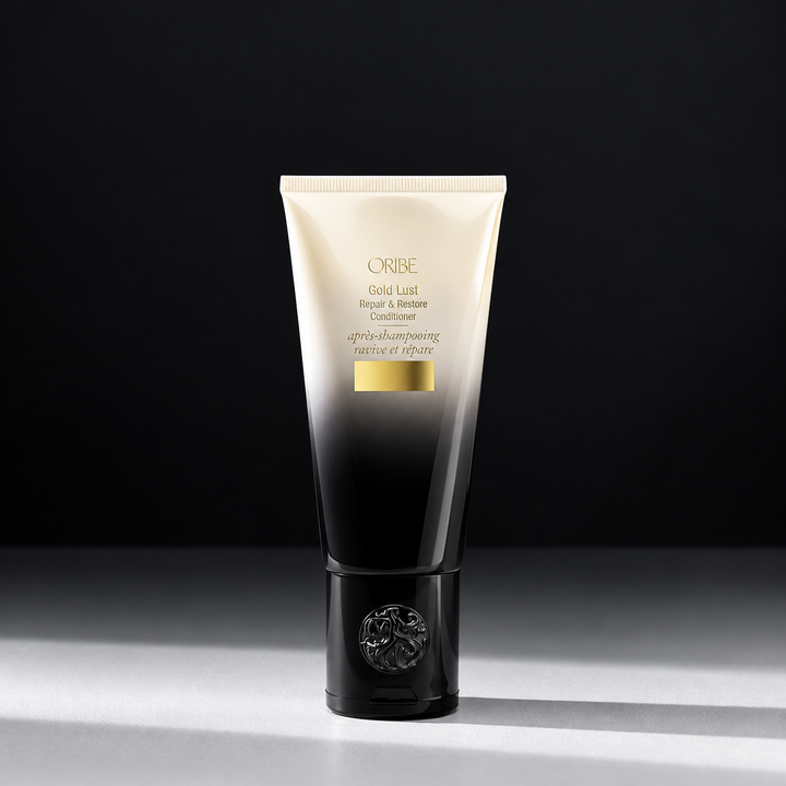 Tube d'après-shampoing ORIBE Gold Lust Repair & Restore, dégradé ivoire et noir, détails dorés et bouchon noir orné du logo, sur fond sombre.