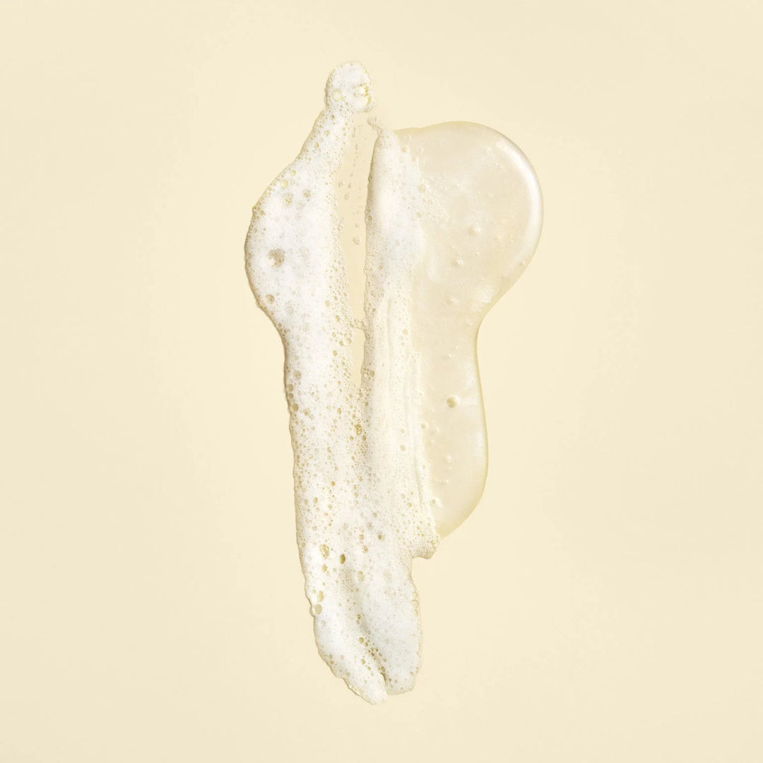 Texture du shampoing ORIBE Gold Lust Repair & Restore, gel translucide légèrement doré avec mousse blanche et fines bulles, étalé sur un fond beige clair.