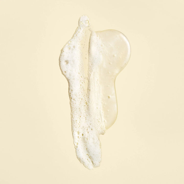 Texture du shampoing ORIBE Gold Lust Repair & Restore, gel translucide légèrement doré avec mousse blanche et fines bulles, étalé sur un fond beige clair.