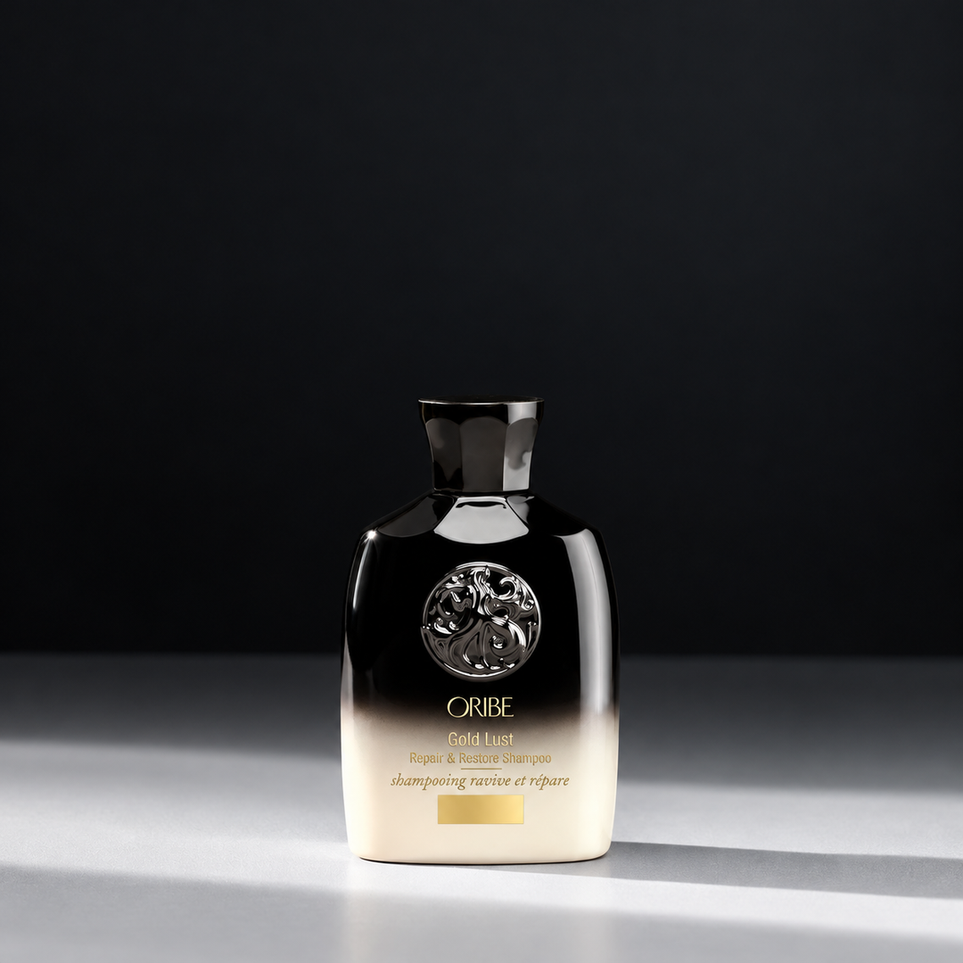 Shampoing réparateur ORIBE Gold Lust Repair & Restore format voyage 75 ml, flacon noir et ivoire avec emblème argenté, inscriptions dorées et bouchon noir, sur fond sombre.