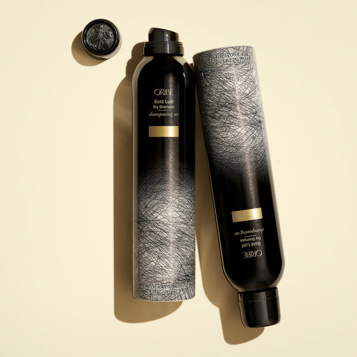 Shampoing sec ORIBE Gold Lust 300 ml présenté avec son flacon aérosol noir et argenté, son capuchon noir retiré et son habillage graphique, sur fond beige clair.
