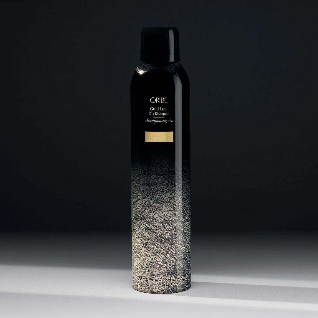 Shampoing sec ORIBE Gold Lust 300 ml, grand flacon aérosol noir dégradé argenté avec motifs graphiques, inscriptions dorées et bouchon noir, photographié sur fond sombre.