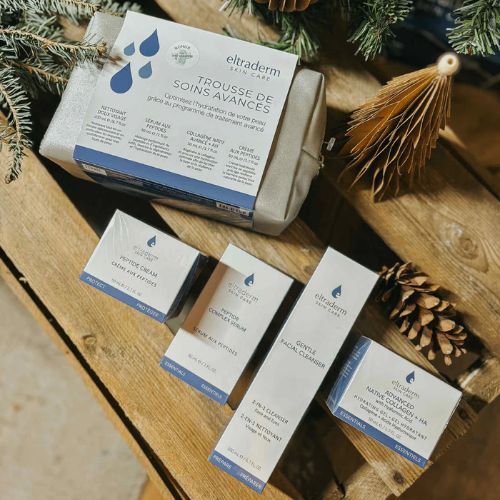 Trousse de soins Eltraderm posée sur une table en bois rustique, accompagnée de cinq produits de soins visage en format boîte et flacon, incluant une crème, un gel nettoyant et un sérum. Emballages blancs et bleus avec logo Eltraderm, décor hivernal avec branches de sapin et pommes de pin. Trousse cosmétique grise incluse. Idéal pour peau hydratée et soins anti-âge.